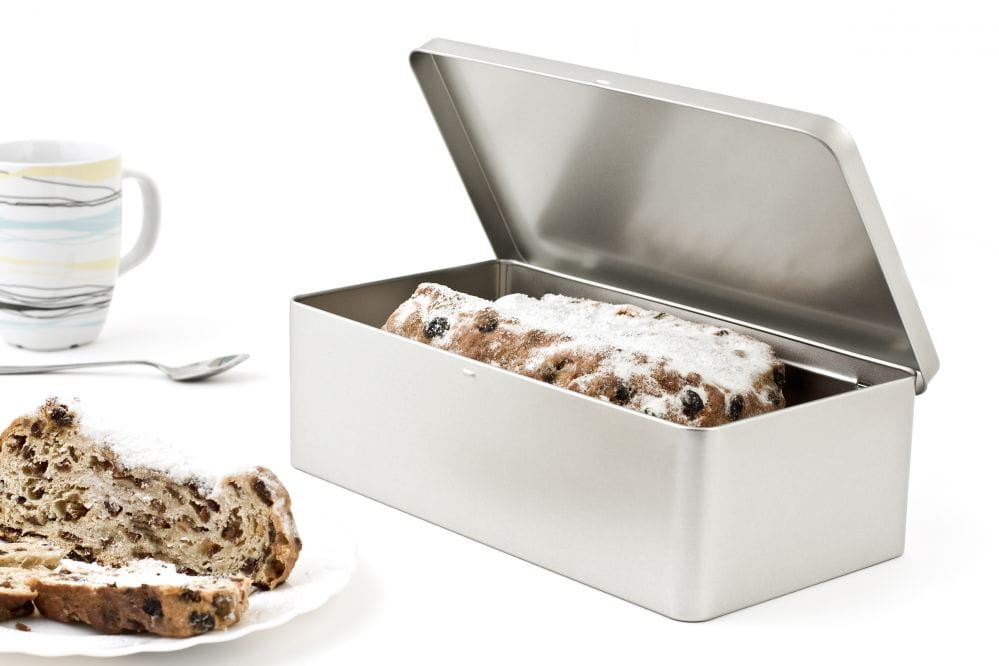 Geöffnete eckige Blechdose mit abgenommenem Stülpdeckel, gefüllt mit Stollen und Puderzucker, auf weißem Hintergrund fotografiert.