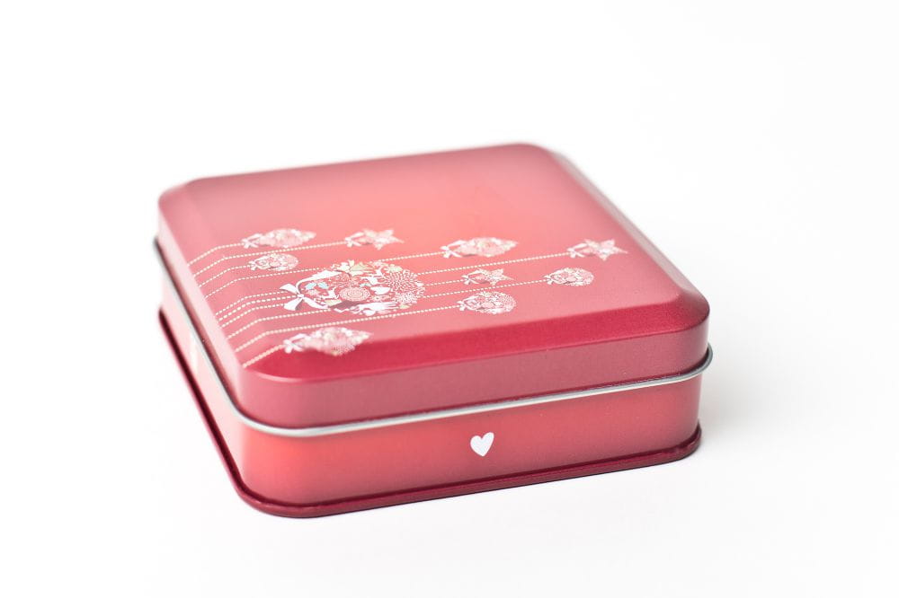 Geschlossene rechteckige Metallbox in Rot mit floralen Verzierungen und Herzmuster auf weißem Hintergrund.